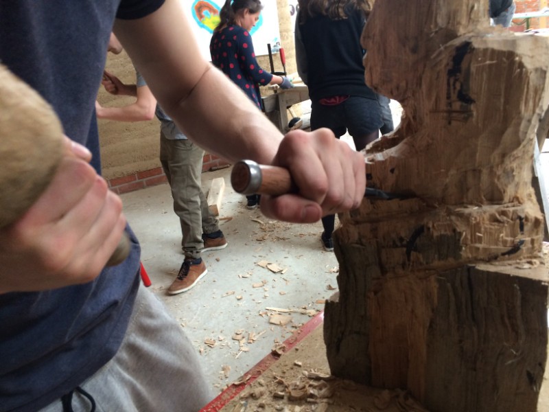 Zu sehen sind zwei Hände, die Hammer und Meißel halten und einen großen Holzblock bearbeiten.
