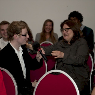 Ein Raum mit roten Stühlen, ein junger Mann mit Mircofon dreht sich zu einer Frau um. Sie und andere Menschen lachen.
