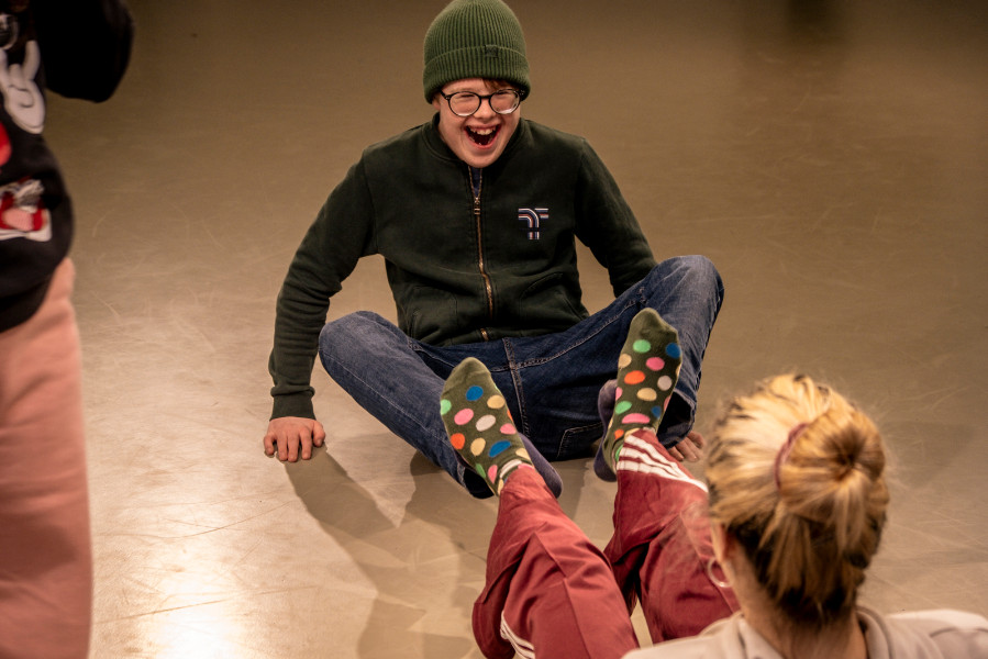 Foto eines Tanzworkshops: Ein lachendes Kind sitzt im Schneidersitz auf dem Boden. Im Vordergrund sind die Beine anderer Kinder sichtbar, darunter eines mit hochgestecktem Haar.