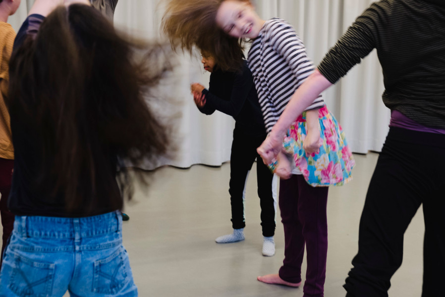 Foto eines Tanzworkshops: Mehrere Kinder tanzen und bewegen sich fröhlich in einem hellen Raum mit weißem Vorhang im Hintergrund. In der Mitte des Bildes steht ein Mädchen, das die Hände anderer Kinder hält, während ihre Haare durch die Bewegung verschwommen wirken.